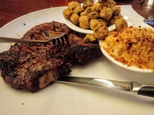 A plate featuring a grilled steak, fried okra, and macaroni and cheese, with a fork and knife beside it.