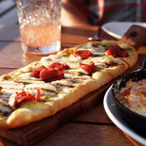 A flatbread topped with mozzarella, sun-dried tomatoes, and pesto, served on a wooden board with a drink in the background.