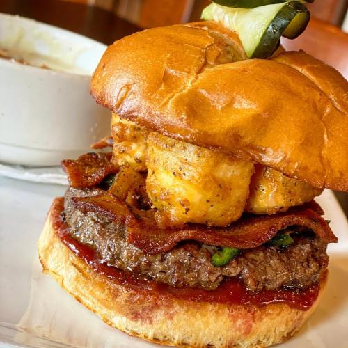 A close-up of a burger with bacon, a crispy topping, and a pickle, served with a bowl of soup on the side.