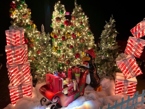 A festive scene with decorated Christmas trees, a sleigh filled with gifts, and a penguin wearing a Santa hat.