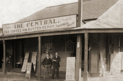 Two men stand outside "The Central" tea, coffee, and butter depot, with vintage signage and a rustic building facade.