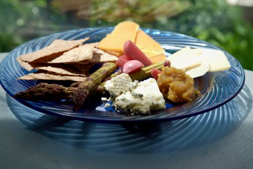 A blue plate with assorted cheeses, crackers, pickled vegetables, and a dollop of chutney, set against a green background.
