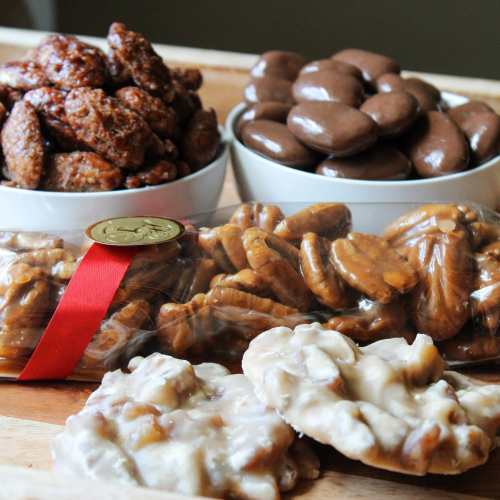 A wooden platter with three bowls of snacks: spiced nuts, chocolate-covered treats, and caramel-coated pecans.