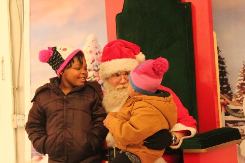 Two children interact with Santa Claus, who is seated on a festive throne, in a cheerful holiday setting.