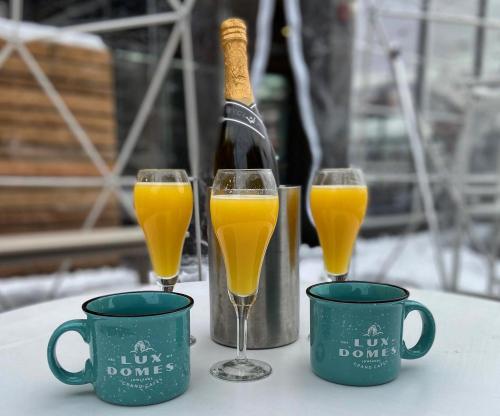 Four glasses of orange mimosa with a bottle of champagne in an ice bucket, alongside two teal mugs on a table.