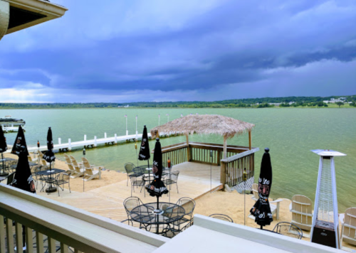 View of a lakeside deck with umbrellas, chairs, and a thatched gazebo under a cloudy sky.