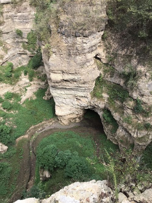 A rocky cliff with a natural arch, surrounded by lush greenery and a winding stream below.