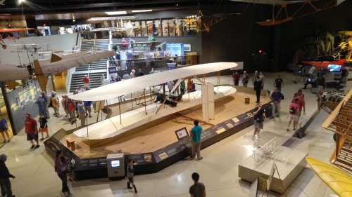 A large model of an early airplane in a museum, with visitors exploring various exhibits in the background.