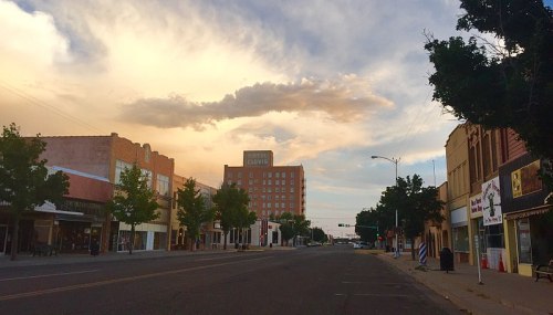 A quiet street scene with trees, storefronts, and a hotel in the background under a cloudy sky at sunset.
