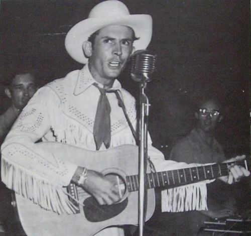 A man in a white cowboy hat and fringed shirt sings into a microphone while playing an acoustic guitar.