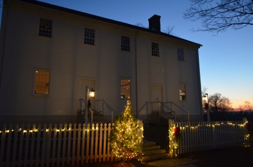 A white house decorated with Christmas lights, featuring a small tree and a picket fence at dusk.