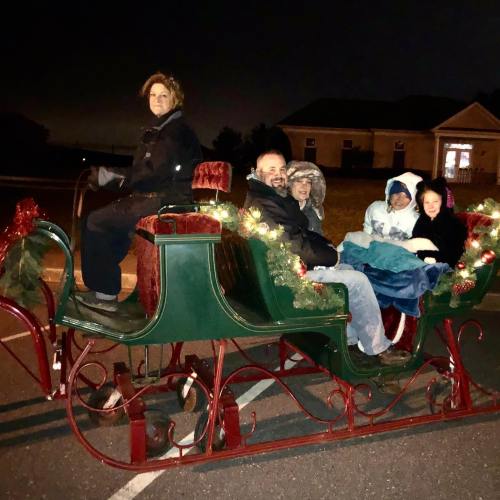 A family sits in a decorated sleigh at night, surrounded by festive lights and greenery.