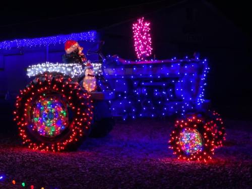 A decorated tractor covered in colorful Christmas lights, with a snowman on top, set against a dark night sky.