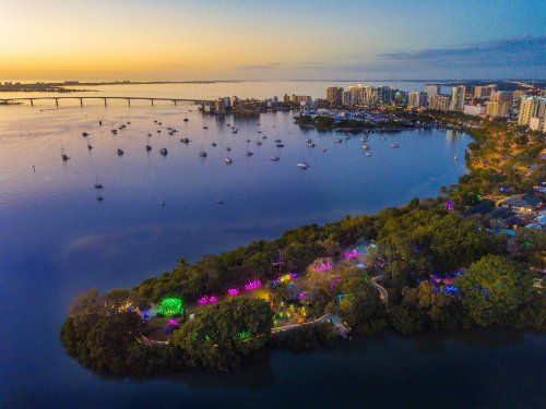 Aerial view of a waterfront city at sunset, featuring colorful lights along the shore and boats in the water.