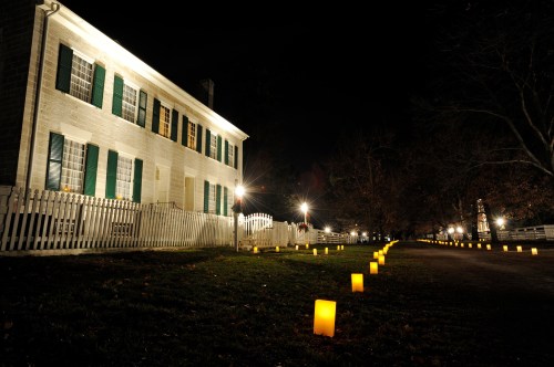 A historic house illuminated at night, with glowing lanterns lining the path and trees in the background.