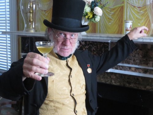 A man in a top hat and formal attire raises a cocktail glass, sitting in a vintage room with decorative wallpaper.