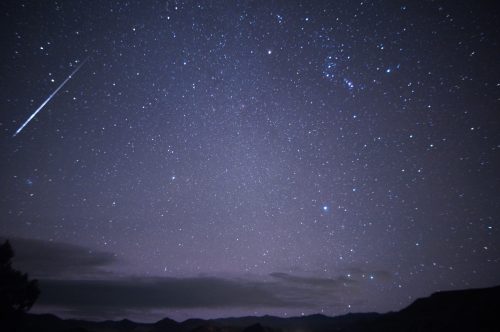 A starry night sky filled with stars, a faint meteor streaking across, and silhouettes of distant mountains below.