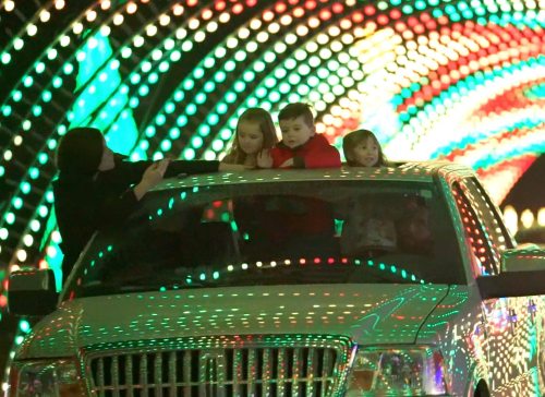 A family enjoys a colorful light display from the roof of a car, with three children smiling and playing.