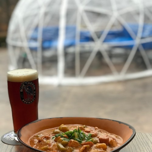 A bowl of stew with vegetables sits on a table next to a pint of beer, with a geodesic dome in the background.