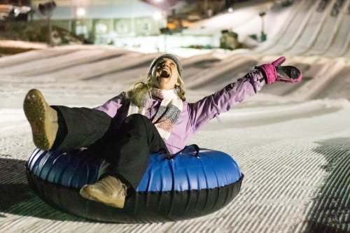 A joyful person in a purple jacket sits on a snow tube, arms raised, enjoying a winter tubing experience.
