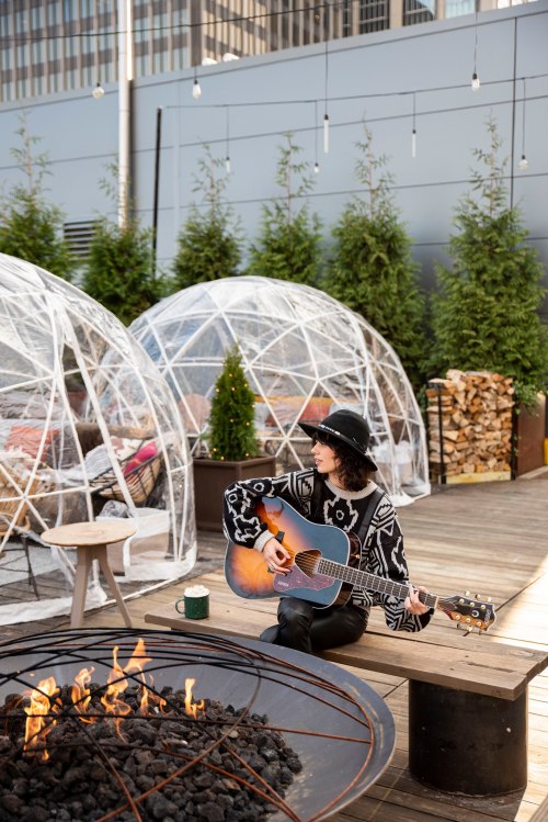 A musician plays guitar near a fire pit, surrounded by cozy igloo-like structures and greenery.