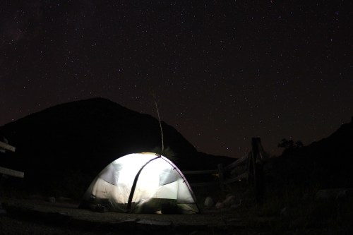 A lit tent under a starry night sky, with mountains silhouetted in the background.