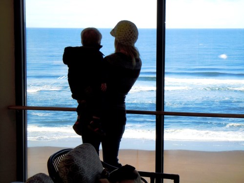 A person holding a child gazes out at the ocean waves through a large window.