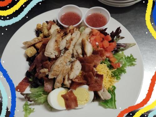 A colorful salad topped with grilled chicken, bacon, cheese, tomatoes, croutons, and two cups of dressing on the side.