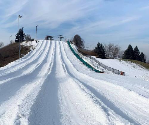 Snow-covered sledding hill with multiple tracks, surrounded by trees and a ski lift in the background under a blue sky.