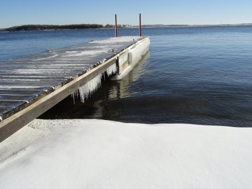 A wooden dock extends over a frozen lake, with icicles hanging and snow covering the shoreline under a clear blue sky.