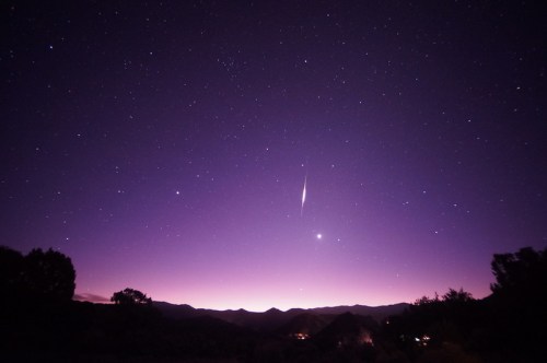 A starry night sky with a purple hue, featuring a bright meteor streaking across the horizon above dark mountains.
