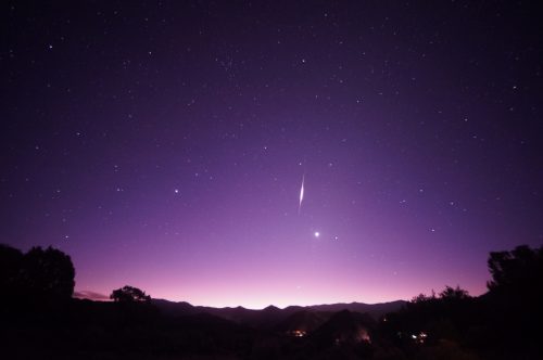 A starry night sky with a purple hue, featuring a bright comet streaking across the horizon above distant mountains.