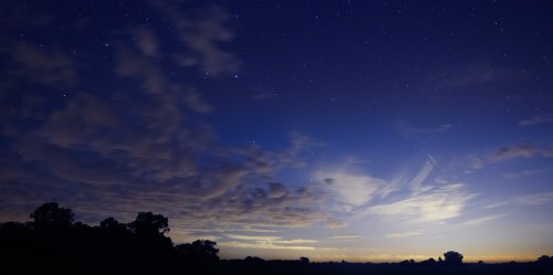 A twilight sky filled with stars and wispy clouds, silhouetted by dark tree outlines on the horizon.