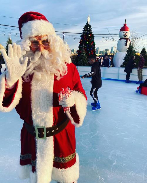 Santa Claus waves while holding candy canes on an ice skating rink, with a snowman and Christmas tree in the background.