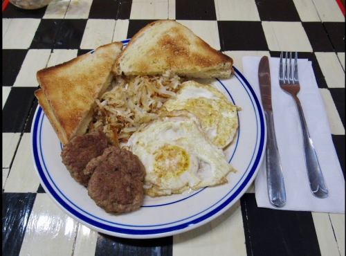 A plate with two fried eggs, hash browns, two sausage patties, and toasted bread on a checkered table.