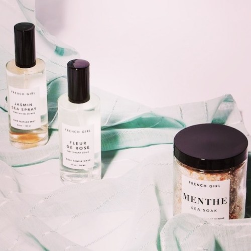 Three beauty products on a light fabric: a jasmine spray, a rose water mist, and a mint sea soak in a jar.