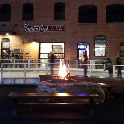 A cozy outdoor plaza with a fire pit, benches, and shops in a brick building, illuminated at night.
