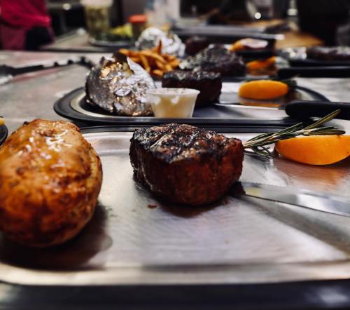 A close-up of a metal tray with grilled steak, baked potato, fries, and garnishes, set in a casual dining environment.