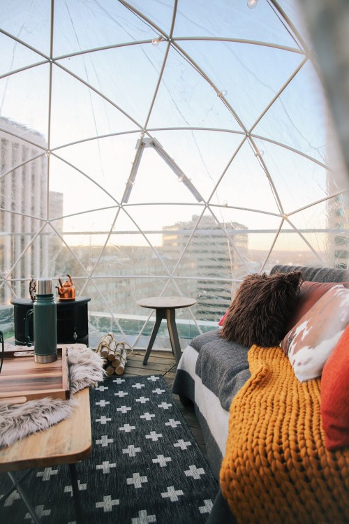 Cozy interior of a geodesic dome with a sofa, table, and city skyline view through transparent walls.