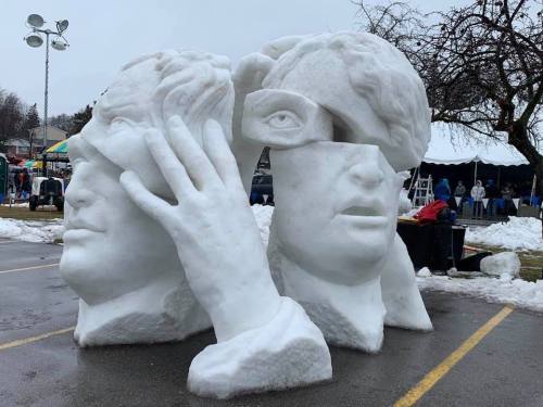 Snow sculpture featuring two stylized human heads with hands and an eye, set in a park during winter.