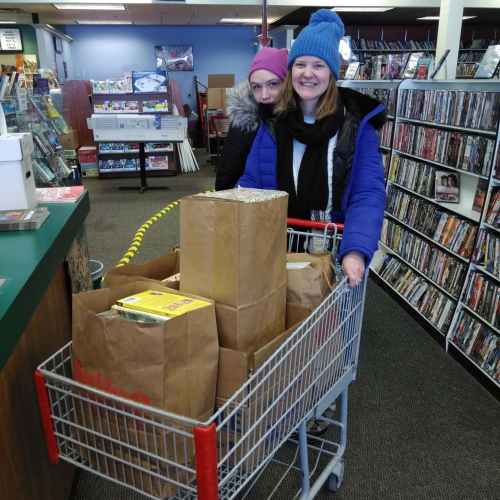 Two women stand behind a shopping cart filled with groceries in a store, with shelves of DVDs in the background.