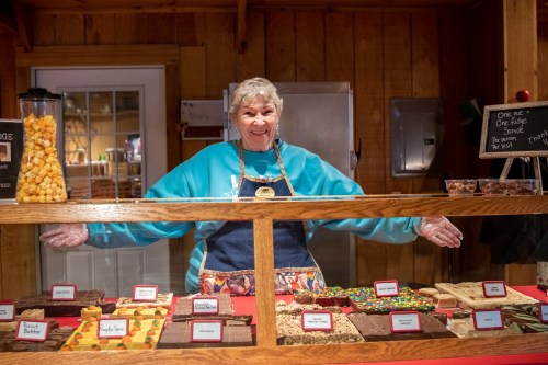 A smiling vendor stands behind a display of colorful fudge and treats in a wooden shop.