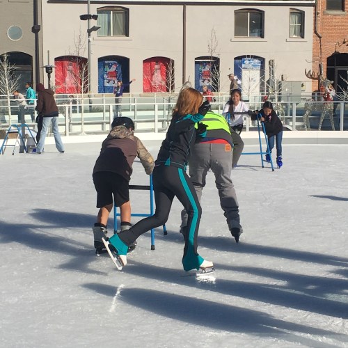 A group of people ice skating on a rink, some using support aids, with buildings in the background.