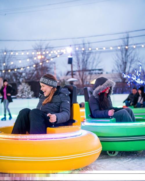 Two women enjoy bumper cars on an ice rink, surrounded by festive lights and other skaters in the background.