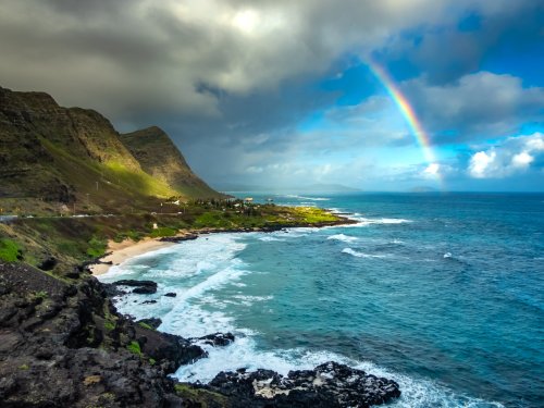 A scenic coastal view featuring a rainbow over the ocean, rocky shores, and lush green hills under a partly cloudy sky.