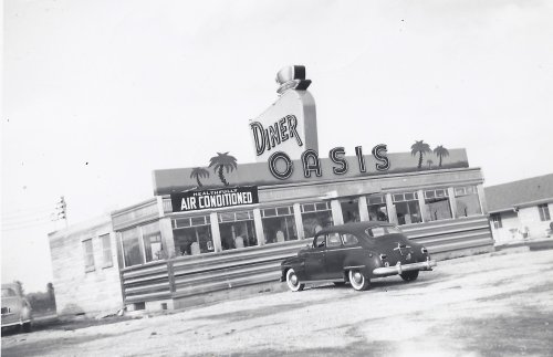 A vintage diner named "Oasis" with palm trees, featuring a classic car parked outside. Black and white photo.