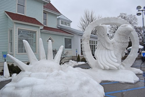 Snow sculptures of a hand and an eagle in front of a light blue building, with some melting snow around.
