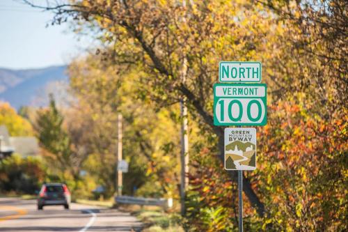 A green road sign for North Vermont 100, surrounded by colorful autumn foliage and a winding road.