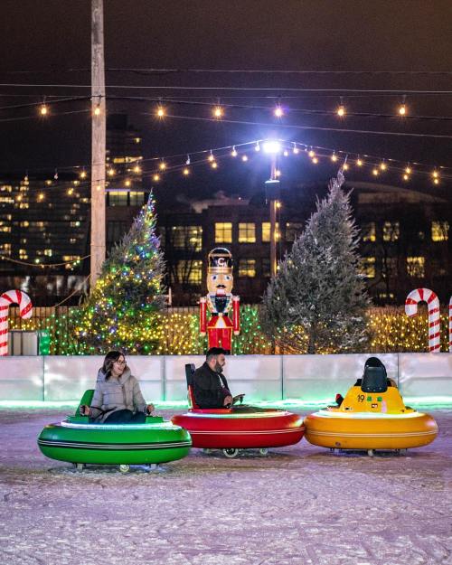 Three people in colorful bumper cars on an ice rink, surrounded by festive lights and holiday decorations.