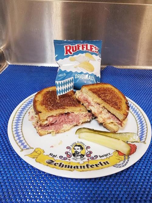 A plate with a grilled sandwich, a pickle, and a bag of Ruffles chips on a blue surface.